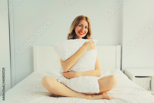 Healthy sleep, happy relaxed young woman hugs her pillow during lying on soft comfortable bed in white bedroom