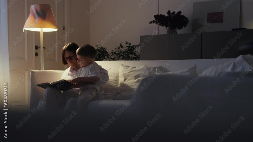 Grandmother and grandson are sitting on a white sofa and reading a book together. A cozy evening at Grandma's. Senior lady and her grandson spend time together. Grandmother and grandson are sitting on a white sofa and reading a book together. A cozy evening at Grandma's. Senior lady and her grandson spend time together.