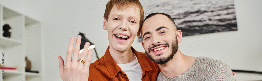 redhead gay man looking at camera and showing wedding ring near overjoyed boyfriend at home, banner. redhead gay man looking at camera and showing wedding ring near overjoyed boyfriend at home, banner.