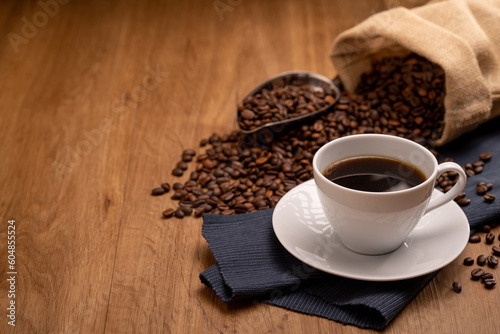 Hot coffee in a white coffee cup and many coffee beans placed around on a wooden table in a warm  light atmosphere  on dark background.