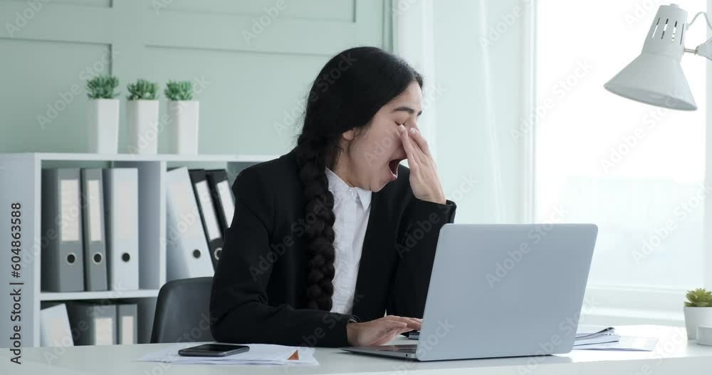 The exhaustion and determination of a tired Indian woman sitting in the office, she continues to work on tasks. As she yawns, she instinctively covers mouth with hand, subtly indicating fatigue. The exhaustion and determination of a tired Indian woman sitting in the office, she continues to work on tasks. As she yawns, she instinctively covers mouth with hand, subtly indicating fatigue.