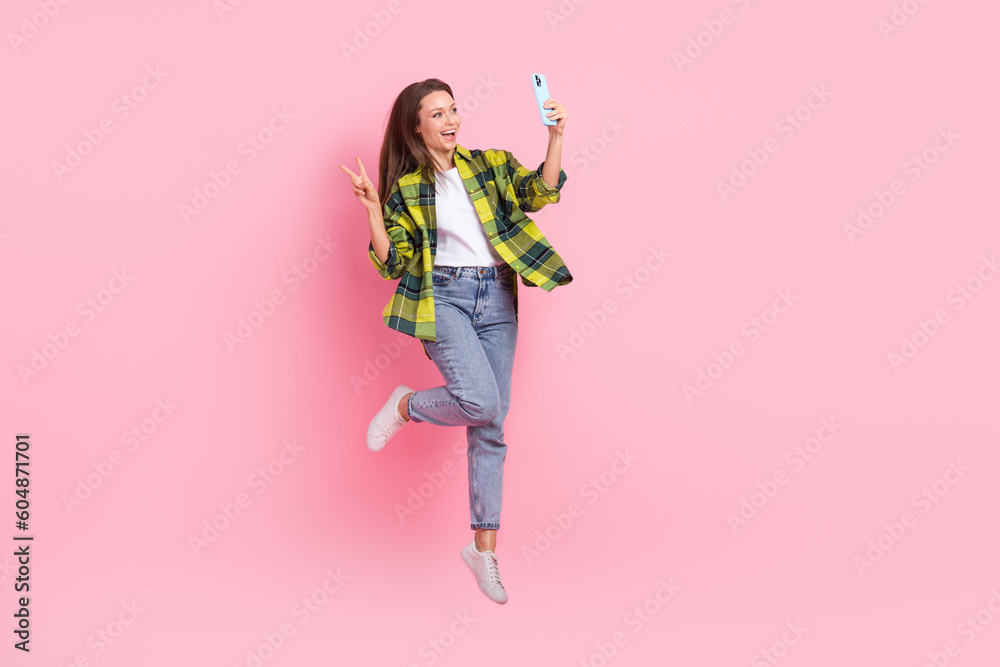 Full length photo of positive woman checkered shirt showing v-sign hold smartphone doing selfie flying isolated on pink color background Full length photo of positive woman checkered shirt showing v-sign hold smartphone doing selfie flying isolated on pink color background