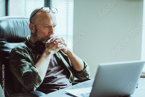 Middle-aged man fully focused on work he looks intently at computer on table. Deep in thought, he reflects on task or problem at hand, demonstrating his analytical and strategic thinking skills. 