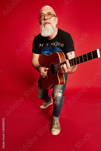 Portrait of beaded, mature man in casual clothes and glaases, playing guitar, picking chords against red studio background. Concept of human emotions, lifestyle, male fashion, hobby