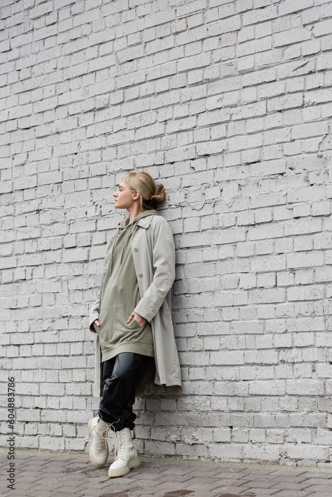 young woman with makeup, blonde hair, bangs, in stylish outfit with long hoodie, coat, black leather pants and beige boots standing with closed eyes near grey brick wall of modern building young woman with makeup, blonde hair, bangs, in stylish outfit with long hoodie, coat, black leather pants and beige boots standing with closed eyes near grey brick wall of modern building