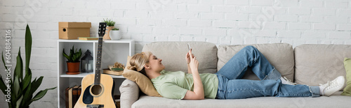 side view of happy woman with blonde and short hair, bangs and eyeglasses using smartphone while resting on comfortable couch near guitar in modern living room with rack and plants, banner