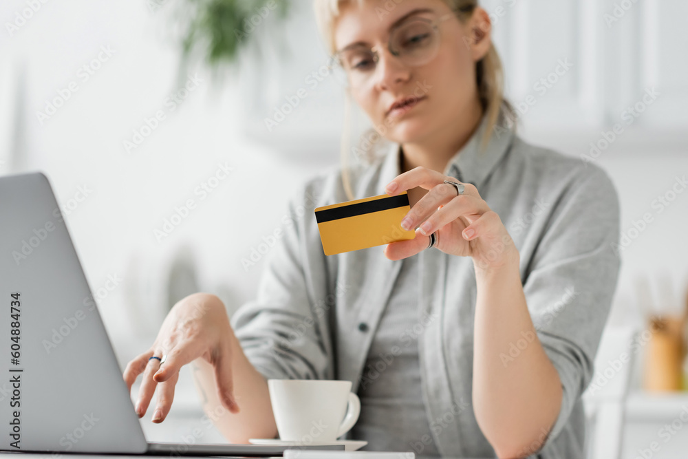 young woman in eyeglasses with tattoo on hand holding credit card, sitting near laptop and cup of coffee on white table, blurred background, work from home, online transactions, technology young woman in eyeglasses with tattoo on hand holding credit card, sitting near laptop and cup of coffee on white table, blurred background, work from home, online transactions, technology