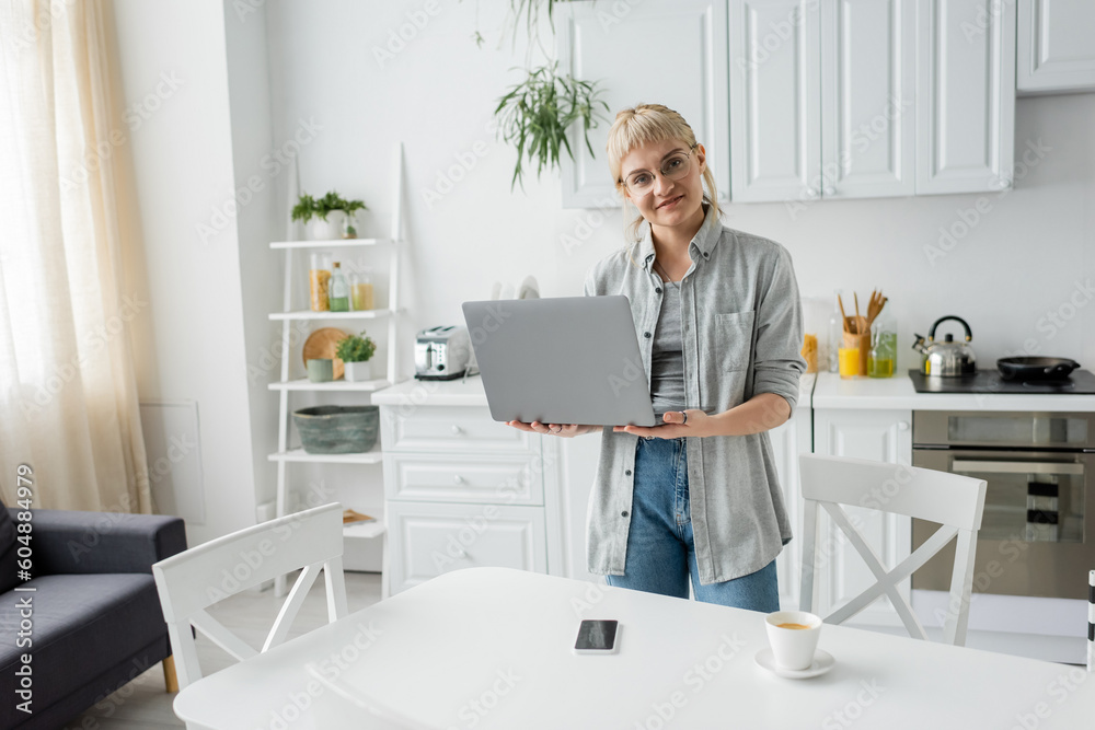 happy young woman in eyeglasses with short hair and bangs holding laptop and looking at camera near cup of coffee and smartphone with blank screen on white table in white and modern kitchen, freelance
