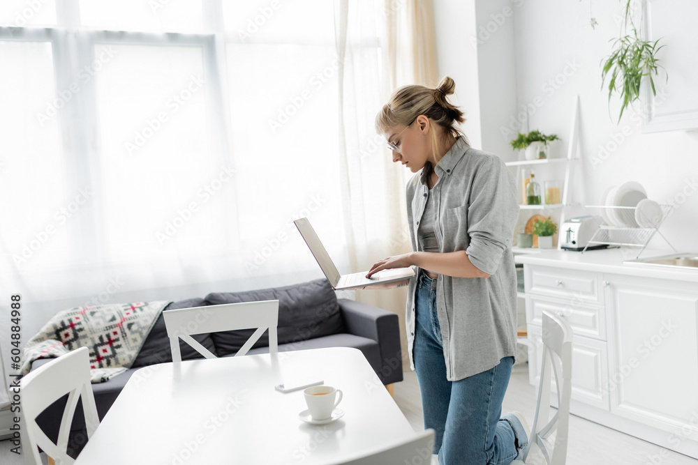 young woman in eyeglasses with short hair and bangs holding laptop near cup of coffee and smartphone on white table around chairs in white and modern kitchen next to sofa, freelancer, remote lifestyle young woman in eyeglasses with short hair and bangs holding laptop near cup of coffee and smartphone on white table around chairs in white and modern kitchen next to sofa, freelancer, remote lifestyle