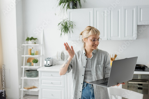 happy young woman in eyeglasses, short hair and bangs holding laptop and waving hand during video call in white and modern kitchen, blurred background, remote lifestyle, freelancer