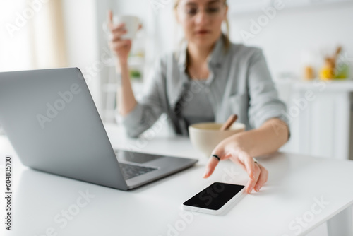 blurred young woman holding cup of coffee near bowl with cornflakes during breakfast while using laptop and reaching smartphone with blank screen in modern kitchen, freelancer, work from home