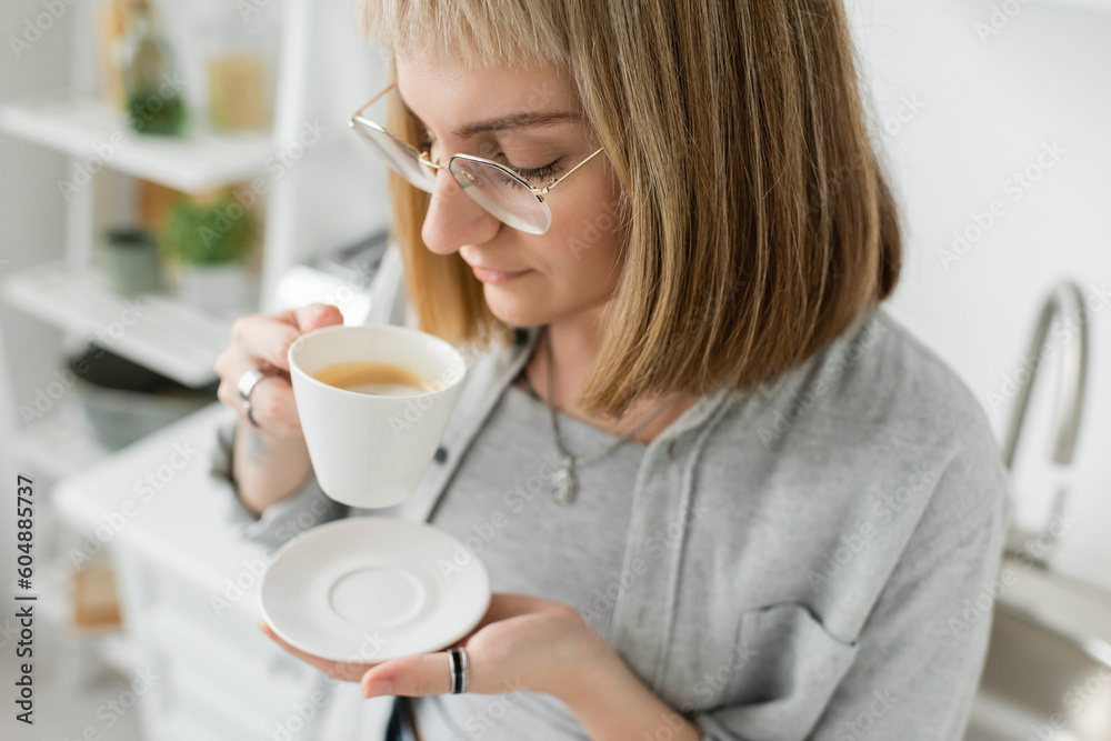 young woman with bangs, eyeglasses and short hair holding cup of morning coffee with saucer and standing in casual grey clothes next to blurred white wall in modern kitchen young woman with bangs, eyeglasses and short hair holding cup of morning coffee with saucer and standing in casual grey clothes next to blurred white wall in modern kitchen