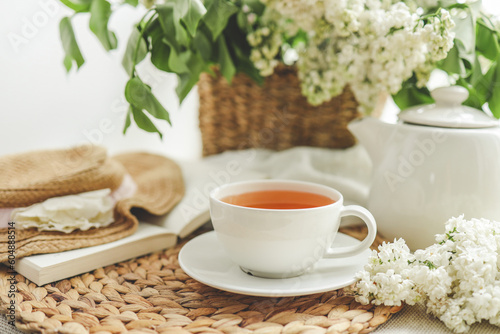 Cup of tea and teapot on windowsill and basket with white lilacs  good morning concept