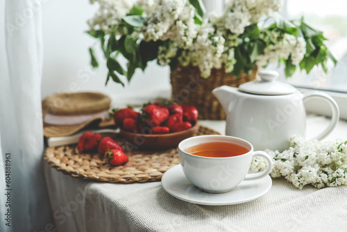 Cup of tea and teapot on windowsill and basket with white lilacs, good morning concept