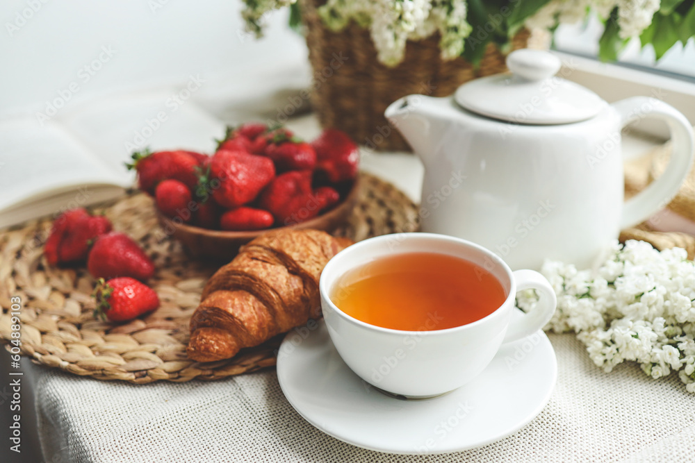 Fototapeta premium Cup of tea, croissant, strawberry and lilac basket, summer morning aesthetic