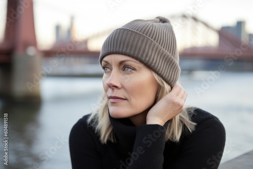 Photography in the style of pensive portraiture of a tender mature woman wearing a warm beanie or knit hat against a picturesque bridge background. With generative AI technology © Markus Schröder