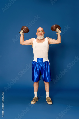 Portrait of bald, bearded, mature man in sportswear posing in vintage boxing gloves against blue studio background. Champion. Concept of human emotions, sport, active lifestyle, sport fashion © master1305
