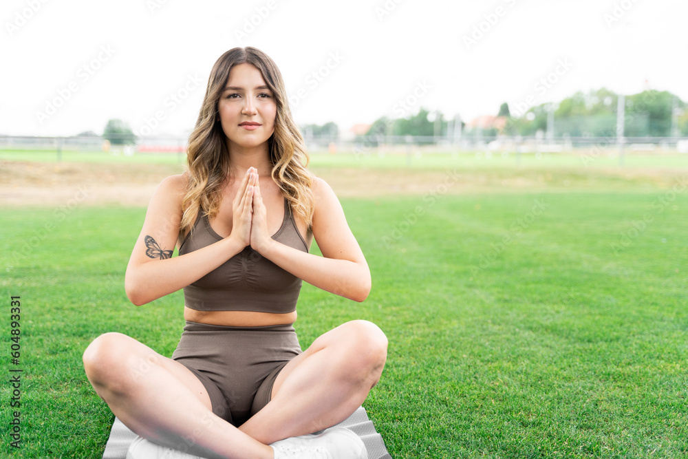 Young caucasian slim fit blond woman sitting in lotus yoga pose on fitness mat in stylish sportwear with hands as in prayer, after workout on green grass meadow near trees in summer park at daytime Young caucasian slim fit blond woman sitting in lotus yoga pose on fitness mat in stylish sportwear with hands as in prayer, after workout on green grass meadow near trees in summer park at daytime