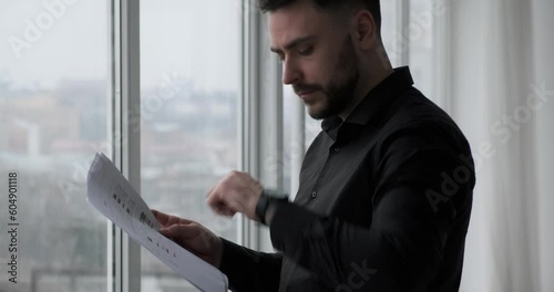 Serious and busy Caucasian businessman in a black suit stands by a window checking documents and glancing at his wristwatch. He appears to be waiting for a business meeting.