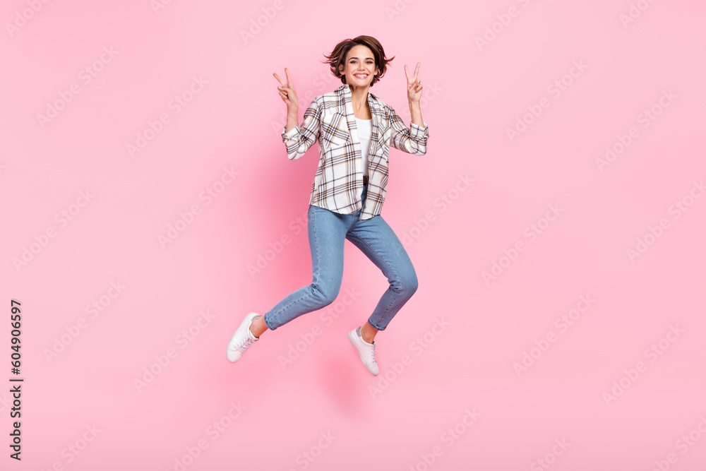 Full body photo of jumping attractive lady double v-sign hello greetings wear plaid shirt with new jeans isolated on pink color background