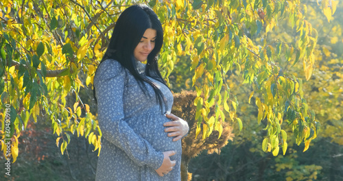 Happy pregnant woman smiling dreaming about baby in sunny autumn park. Pregnant mother strokes, touches belly on nature background of yellow trees foliage. Bright natural sun light day in fall forest