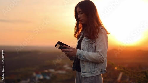 Beautiful young woman reading the bible while standing on top of a hill with a beautiful view at sunset, admiring the view and nature, relaxing alone with thoughts, slow motion