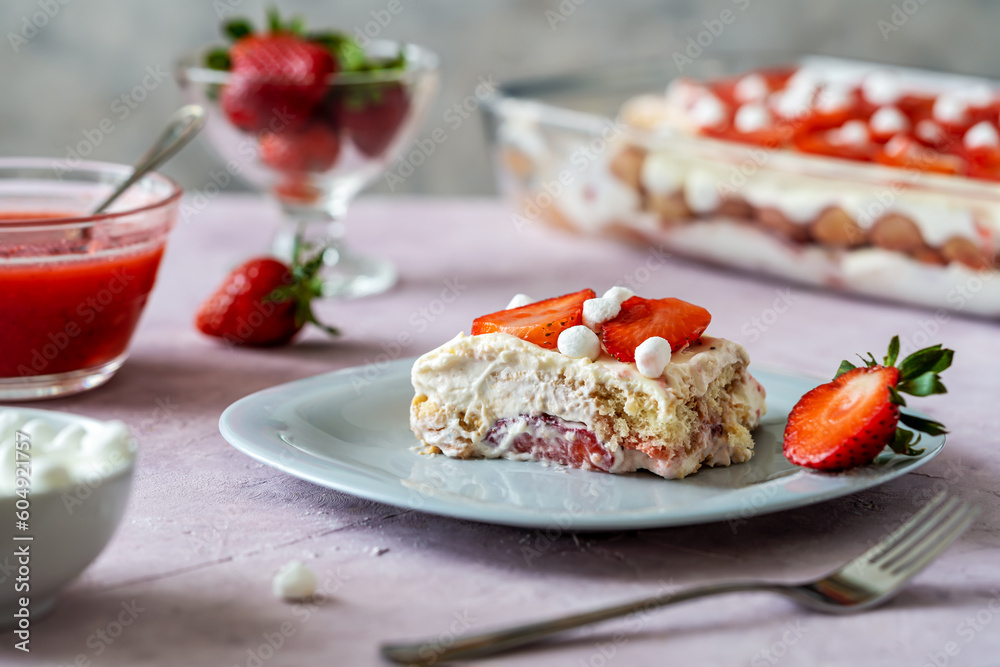 Italian traditional dessert tiramisu with strawberries. One piece of cake on a plate closeup, whole cake and strawberry juice backside, rose and grey concrete background
