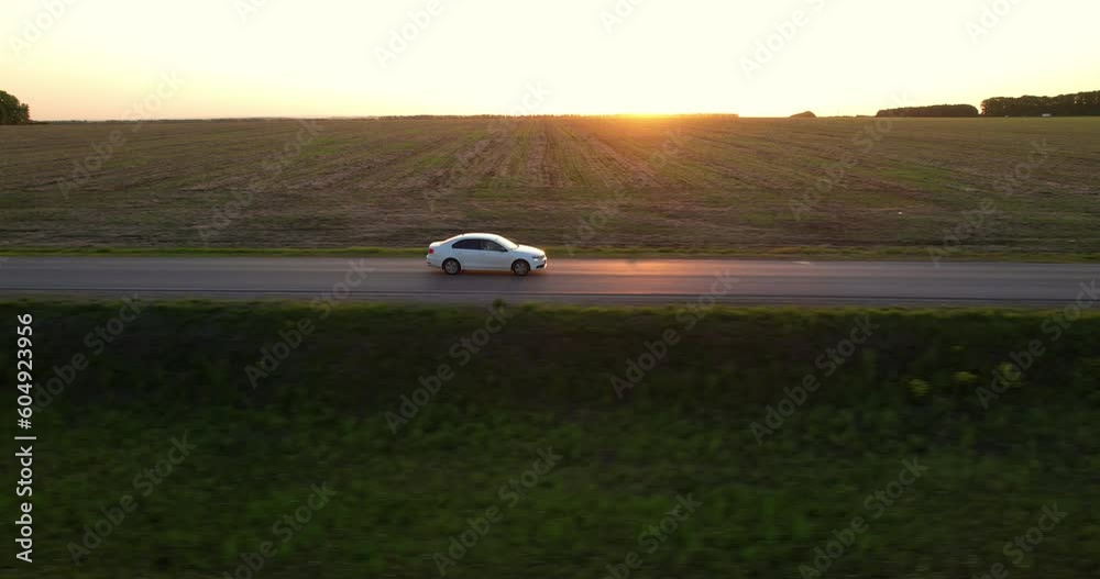 A white car is driving straight on an asphalt road during sunset, aerial shot. Side view. Rays of the setting sun. Sunlight is reflected on the surface of the asphalt road. 