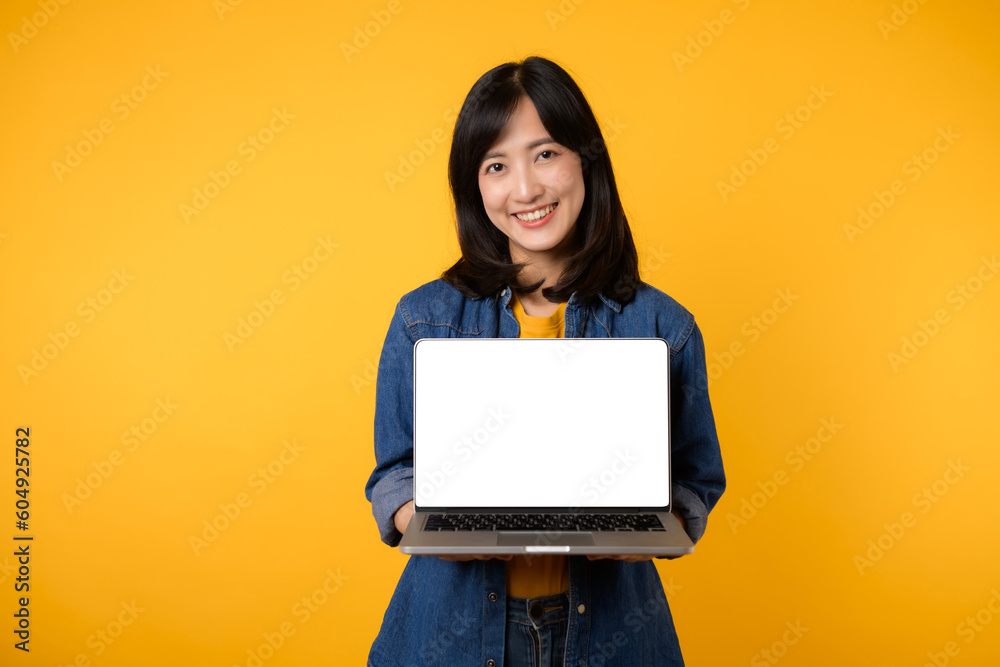 Naklejka premium portrait young happy woman wearing yellow t-shirt and denim shirt holding laptop and point finger to screen isolated on yellow studio background. business technology application communication concept.