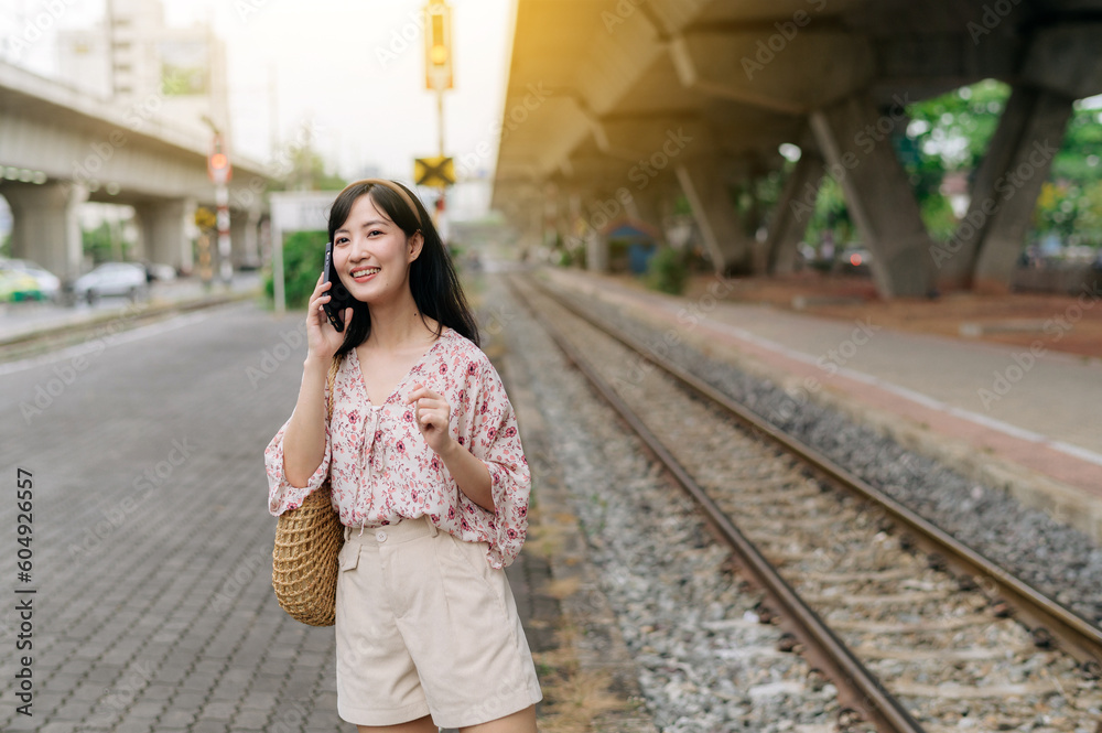 Fototapeta premium Asian young woman traveler with weaving basket using a mobile phone beside railway train station in Bangkok. Journey trip lifestyle, world travel explorer or Asia summer tourism concept.