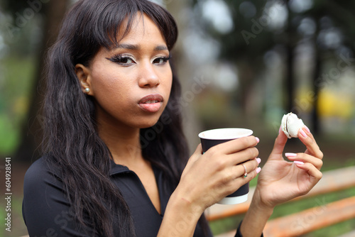 A brunette woman is resting in the park With a cup of coffee and macarons.