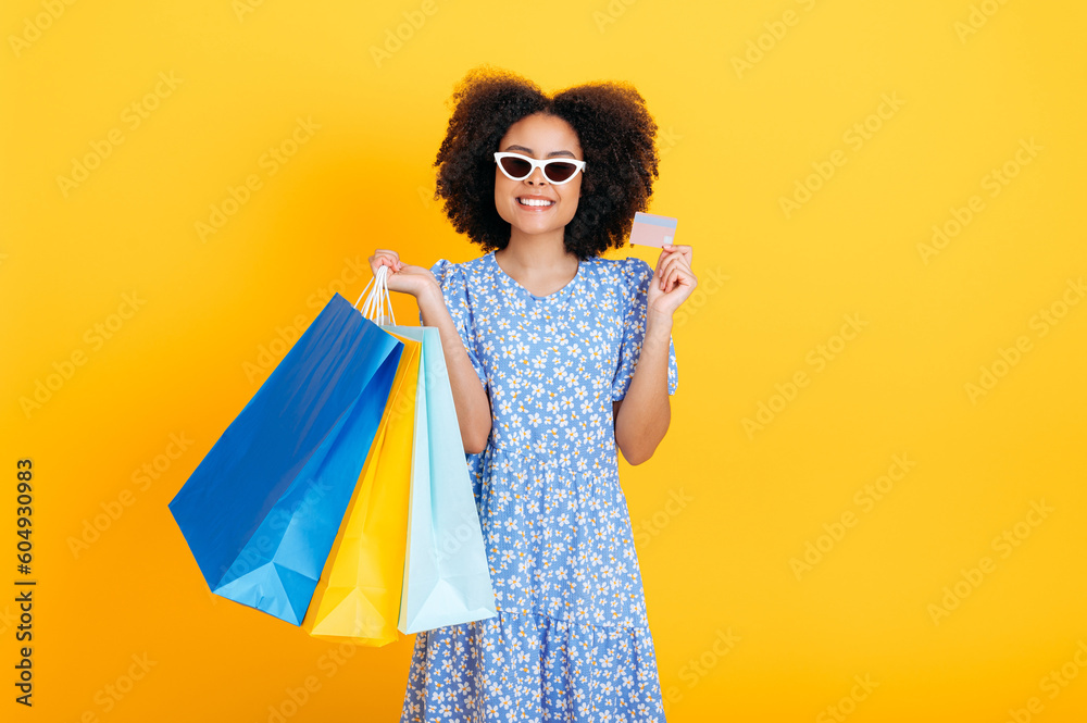 Photo of charming smiling positive african american or brazilian young woman wearing blue sundress, with sunglasses, holding paper shopping bags and credit card, stand on isolated yellow background Photo of charming smiling positive african american or brazilian young woman wearing blue sundress, with sunglasses, holding paper shopping bags and credit card, stand on isolated yellow background