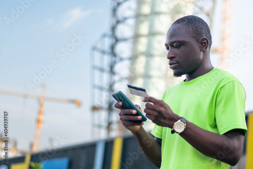 A man in a green shirt looking at a cell phone and holding credit card