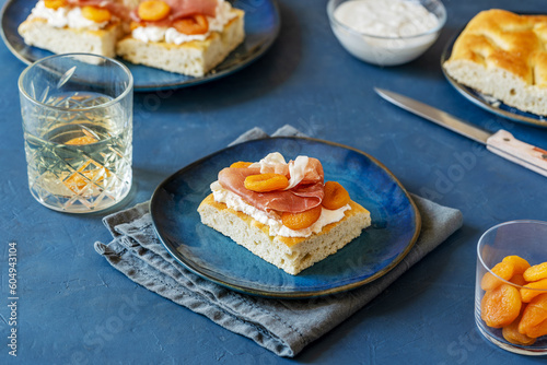 Focaccia with prosciutto, soft cheese stracciatella and dry apricots on a blue plate, grey napkin, drink, ingredients and others sandwiches backside over dark blue background
