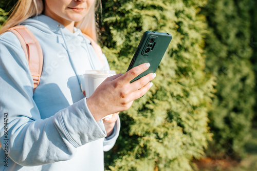 Close-up of caucasian woman in casual clothes using smartphone outdoors. Woman holding mobile phone and cup of coffee in nature