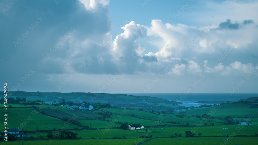Cloudy sky over the green rolling fields of southern Ireland. Landscape of the southern coast of Ireland with blue sky and clouds. Green grass field under white clouds. Cloudy sky over the green rolling fields of southern Ireland. Landscape of the southern coast of Ireland with blue sky and clouds. Green grass field under white clouds.