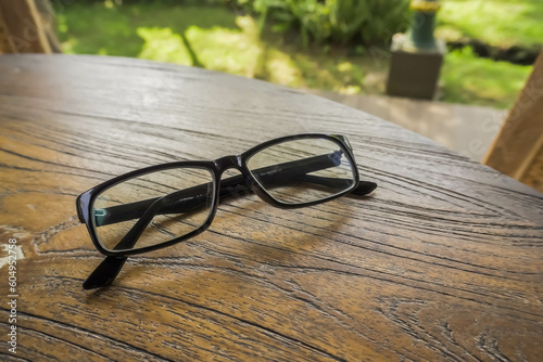 Black framed glasses lied on a wooden table with bokeh background. Side view daylight shade during the day. Concept for working, task, tired eyes, display monitor radiation, job done, smart person.