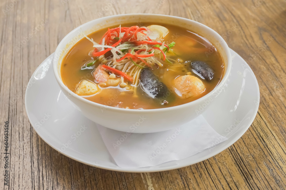 Tom Yum in white bowl isolated on wooden table with beautiful garnish. Tom Yum is a type of hot and sour Thai soup, usually cooked with shrimp or prawn with lots of herbs and spices. Tom Yum in white bowl isolated on wooden table with beautiful garnish. Tom Yum is a type of hot and sour Thai soup, usually cooked with shrimp or prawn with lots of herbs and spices.