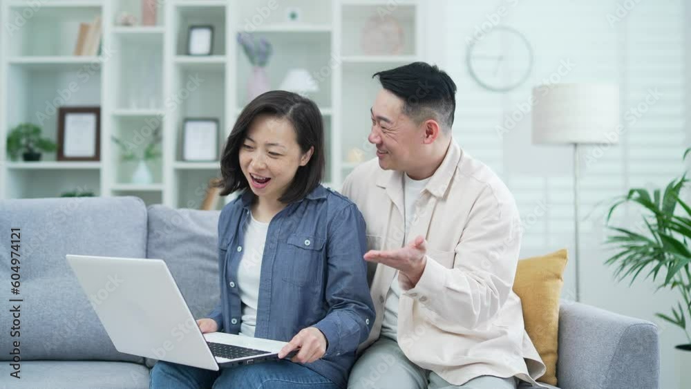 Happy asian family couple talking on video call using laptop, waving hands while sitting on sofa in living room at home. Loving wife and husband cuddles, smiles, have a conversation looking at camera Happy asian family couple talking on video call using laptop, waving hands while sitting on sofa in living room at home. Loving wife and husband cuddles, smiles, have a conversation looking at camera