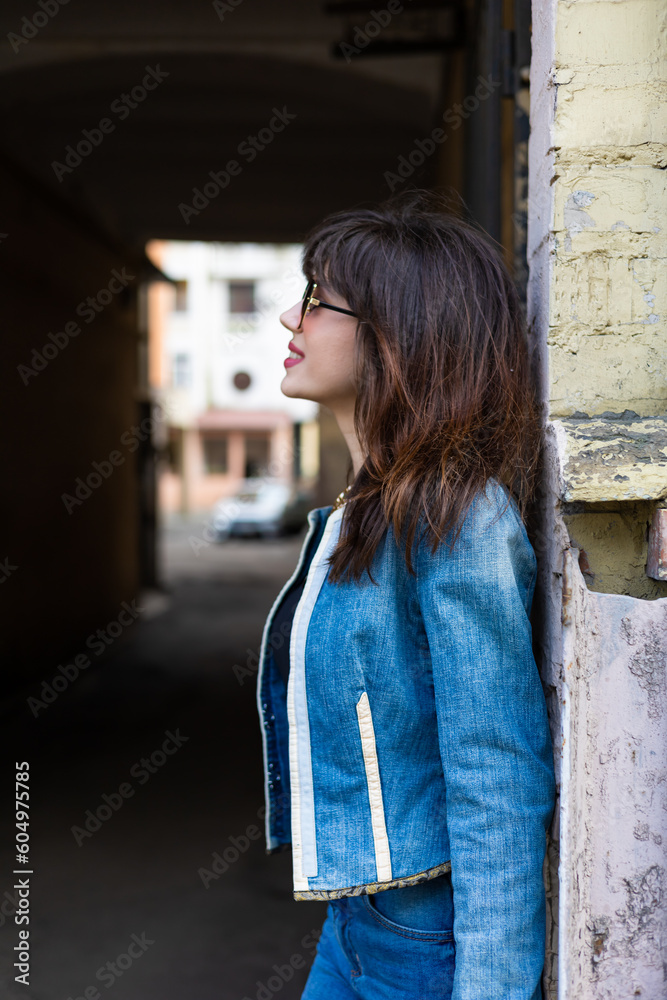 Cute young girl with dark wavy hairstyle and bright makeup, jeans suit, wearing sunglasses posing in arch in city street. 