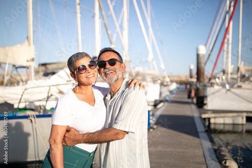 Cheerful Mature Spouses Embracing Posing At Marina With Yachts Outside