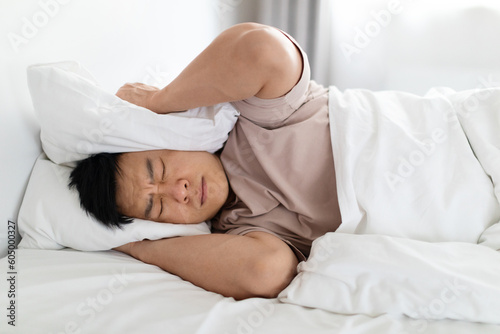 Irritated korean man lying in bed, covering head with pillow