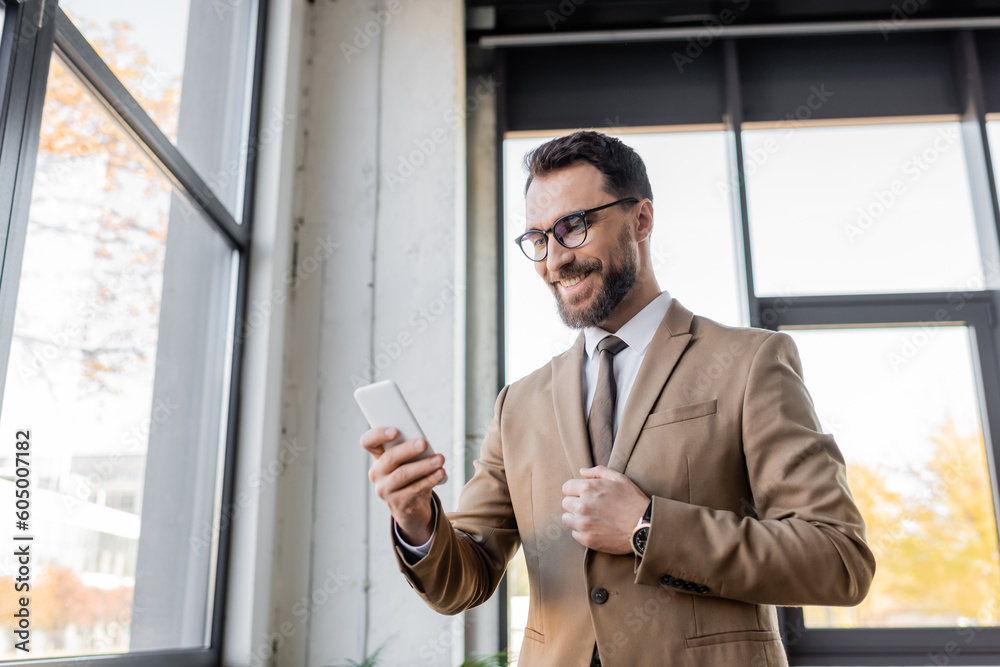 Fototapeta premium charismatic and cheerful entrepreneur in trendy business attire such as beige blazer and eyeglasses standing near windows in office and looking at mobile phone