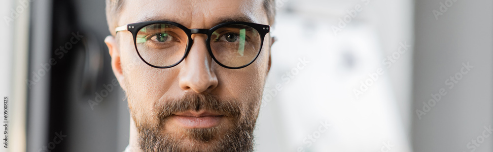 portrait of good looking and ambitious businessman with beard and mustache wearing fashionable eyeglasses and looking at camera in blurred office, banner