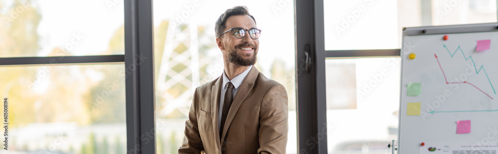Fototapeta premium successful bearded businessman in stylish eyeglasses, beige blazer and tie looking away and smiling near flip chart with graphs and sticky notes in office with large windows, banner