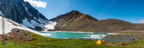Back country camping area in northern arctic Canada during summer time with panoramic view over glacial lake, yellow tent, huge mountains, snow, ice, green grass. 