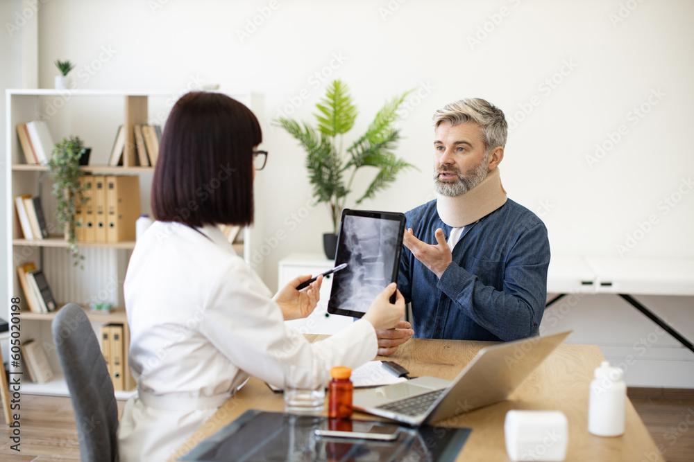 Mature bearded man in C collar talking to female physician in white coat pointing at tablet with spine scan on screen. Worried patient explaining health condition to medical person in consulting room. Mature bearded man in C collar talking to female physician in white coat pointing at tablet with spine scan on screen. Worried patient explaining health condition to medical person in consulting room.