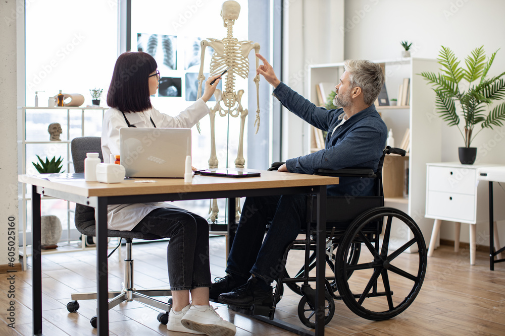 Full length view of female physician and man with disability examining skeleton model while staying in doctor's office. Attentive adults analyzing spinal cords for better awareness of pain source. Full length view of female physician and man with disability examining skeleton model while staying in doctor's office. Attentive adults analyzing spinal cords for better awareness of pain source.