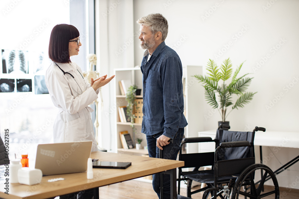 Smiling female with stethoscope clapping while happy man standing on his feet after using wheelchair. Cheerful doctor in consulting room congratulating patient with regaining mobility after accident. Smiling female with stethoscope clapping while happy man standing on his feet after using wheelchair. Cheerful doctor in consulting room congratulating patient with regaining mobility after accident.