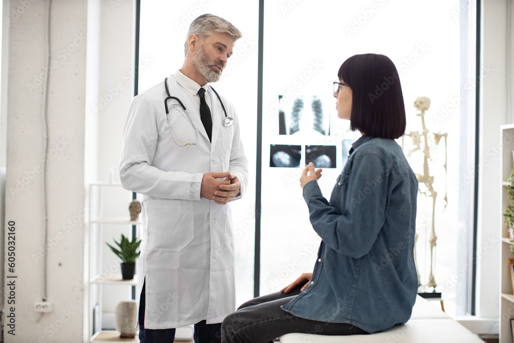 Obraz premium Pretty bespectacled lady in everyday wear having conversation with gray-haired caucasian male in doctor's coat. Healthcare practitioner offering professional treatment for illness in medical center.